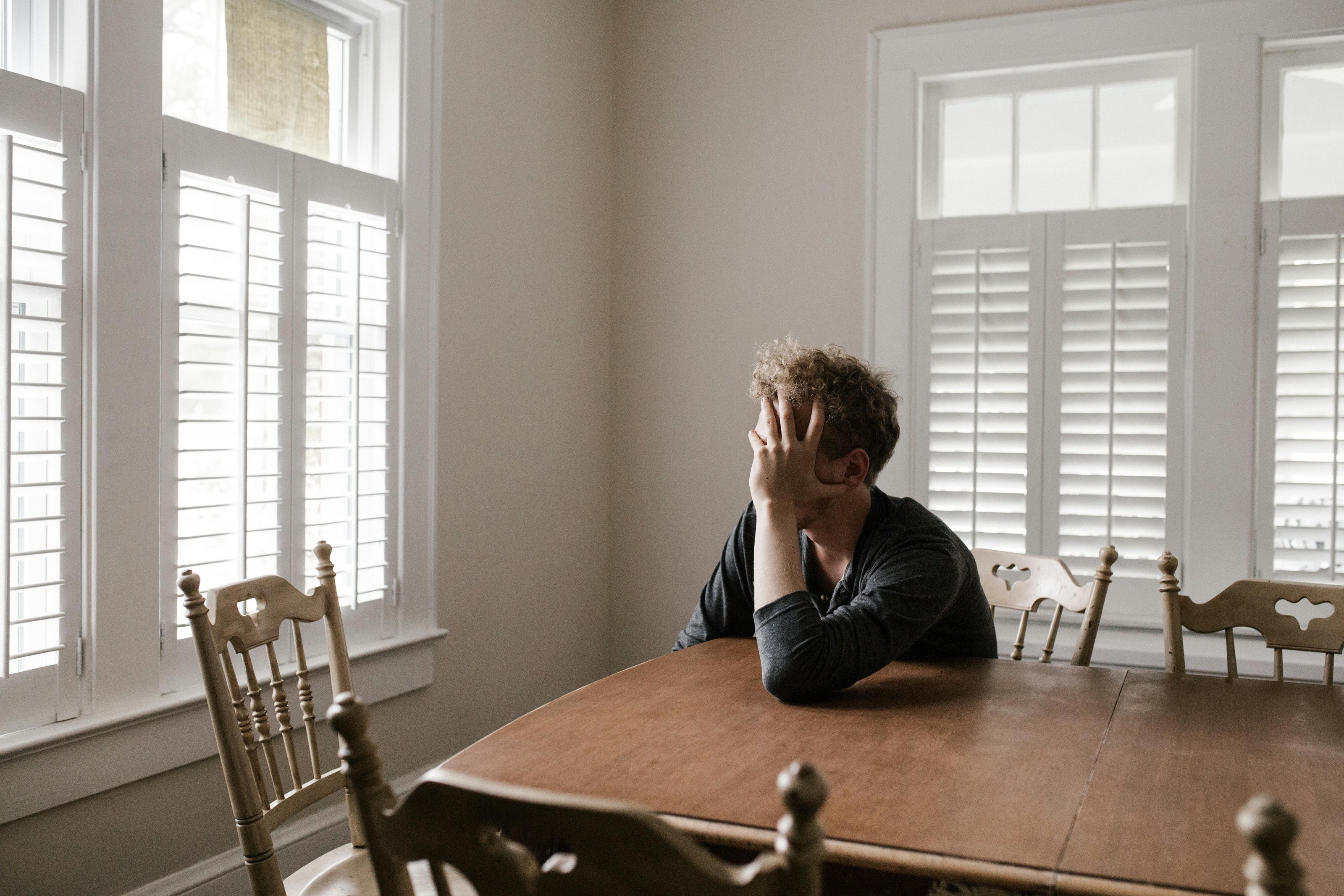 Person sitting alone at a wooden dining table in a room with large windows.